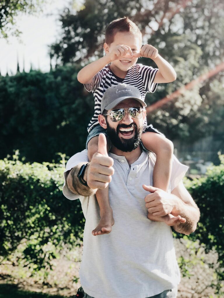 Father giving son a ride on shoulders, both laughing and smiling in a park setting.